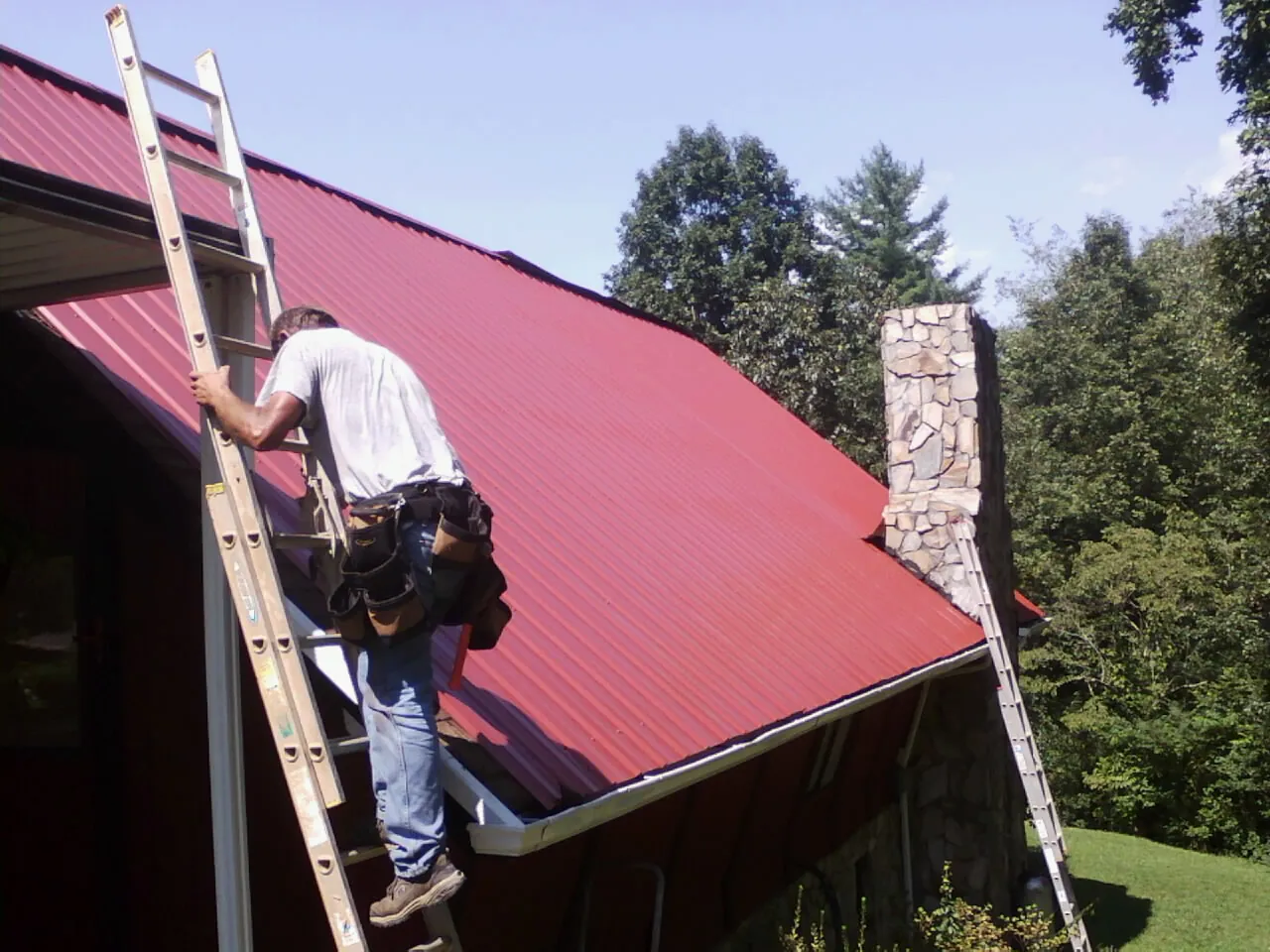 Roofer on a ladder beside a red metal roof and stone chimney — Soffit & Fascia Repair by Roofing Brothers in Aneta